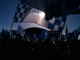 Blue and white flag waving over crowd of people, stage in the distance.