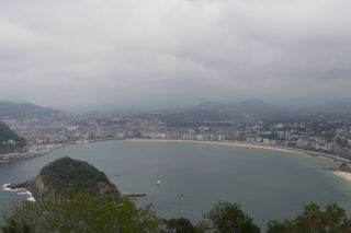 The view from the top of Monte Igueldo towards La Concha in San Sebastián. The shell shaped beach has little activity in the water, the sky is overcast but not gloomy, and there is a light fog over the buildings in the distance.